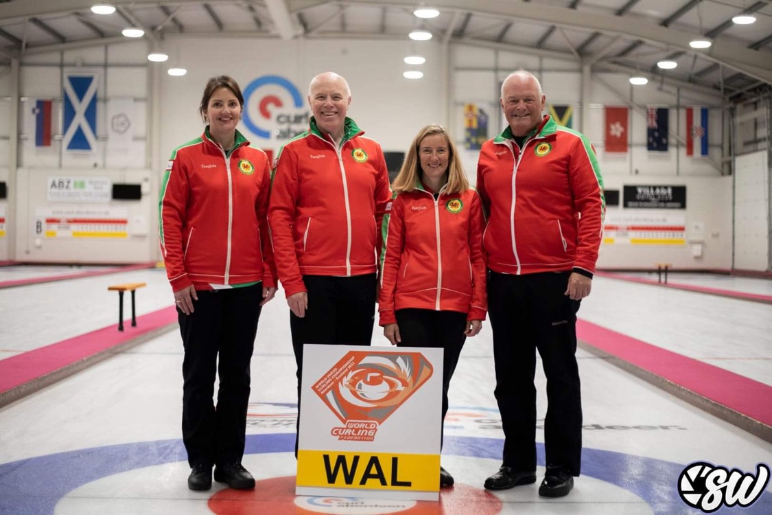 A photo of two men and two women stood in a line on a curling rink. They are all wearing red jackets and black trousers, with a sign for Wales in front of them.